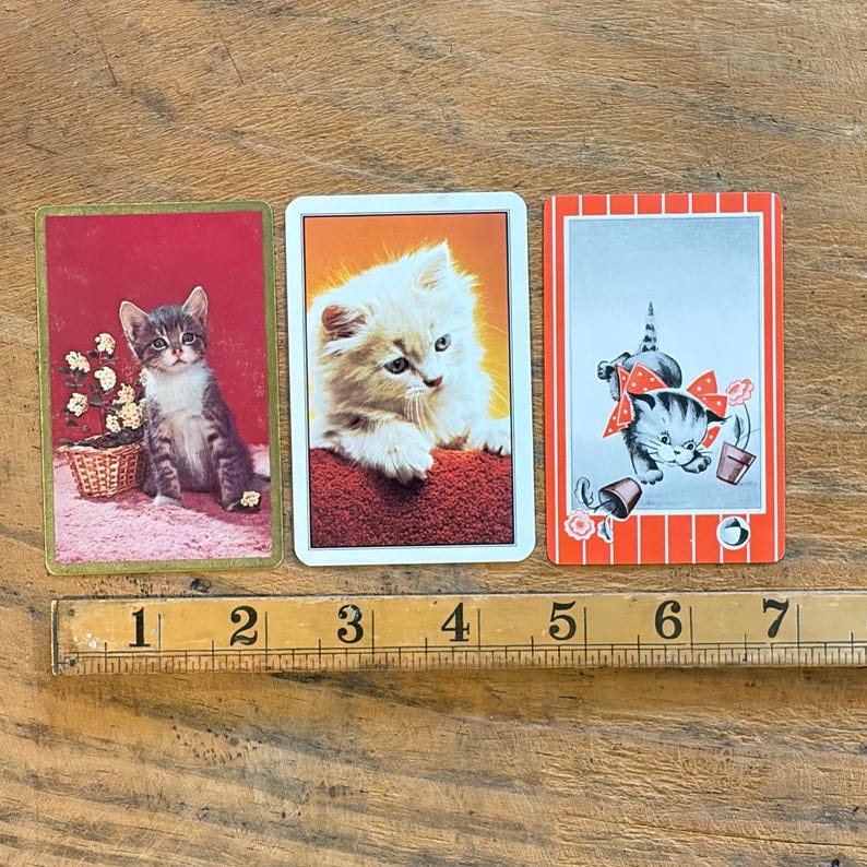 May include: Three rectangular playing cards featuring kitten illustrations, positioned above a wooden ruler measuring 7 inches. The left card shows a gray tabby kitten, the center card a fluffy white kitten, and the right card a cartoon kitten with a red bow.