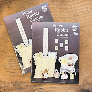 以下が含まれることがあります： Green Apple Co.の「Peter Rabbit Counts」というタイトルの全く同じクラフトパターンブック2冊。ダークブラウンの表紙には、縦型の数字バナー、ウサギのモチーフが描かれた黄色いブランケット、白い籐の椅子、小さな額縁に入ったアートのイラストが掲載されています。