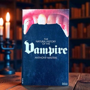 May include: A vintage book titled "The Natural History of the Vampire" by Anthony Masters. The cover shows vampire fangs with blood and the title in a gothic font against a dark blue background. The book is on a wooden surface.