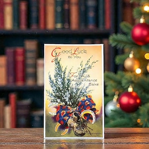 May include: Vintage postcard with the text "Good Luck to You" and "For Auld Acquaintance Sake." The image features a sprig of greenery, a plaid bow, a horseshoe, and a boot. The background includes a bookshelf and Christmas decorations.