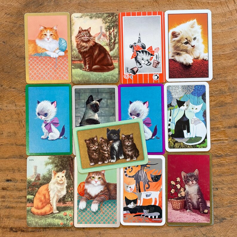 May include: A collection of twelve rectangular playing cards featuring various cat illustrations, including kittens with yarn, a Siamese cat, and artistic feline portraits. The cards display diverse color palettes and artistic styles.