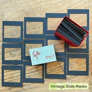 May include: Vintage slide masks and a box of slides are arranged on a wooden surface. The slide masks are black, rectangular, and surround a central opening. A red box contains a stack of black slides. A small, light blue box with red text reads "Sytec SEVEN 0 21 x 31 slides."