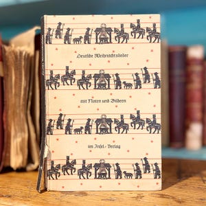 May include: A hardcover book titled Deutsche Weihnachtslieder mit Noten und Bildern im Insel-Verlag. The cream-colored cover features repeating horizontal rows of black illustrations depicting shepherds, sheep, and kings on horseback.