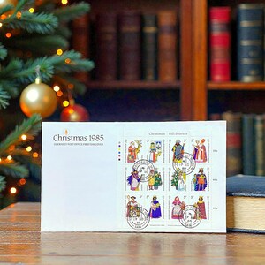 Könnte beinhalten: Ein weißer Umschlag mit dem Text "Christmas 1985" und einer Reihe farbenfroher Briefmarken mit Darstellungen der Geschenkebringer. Die Briefmarken stammen vom Guernsey Post Office First Day Cover.