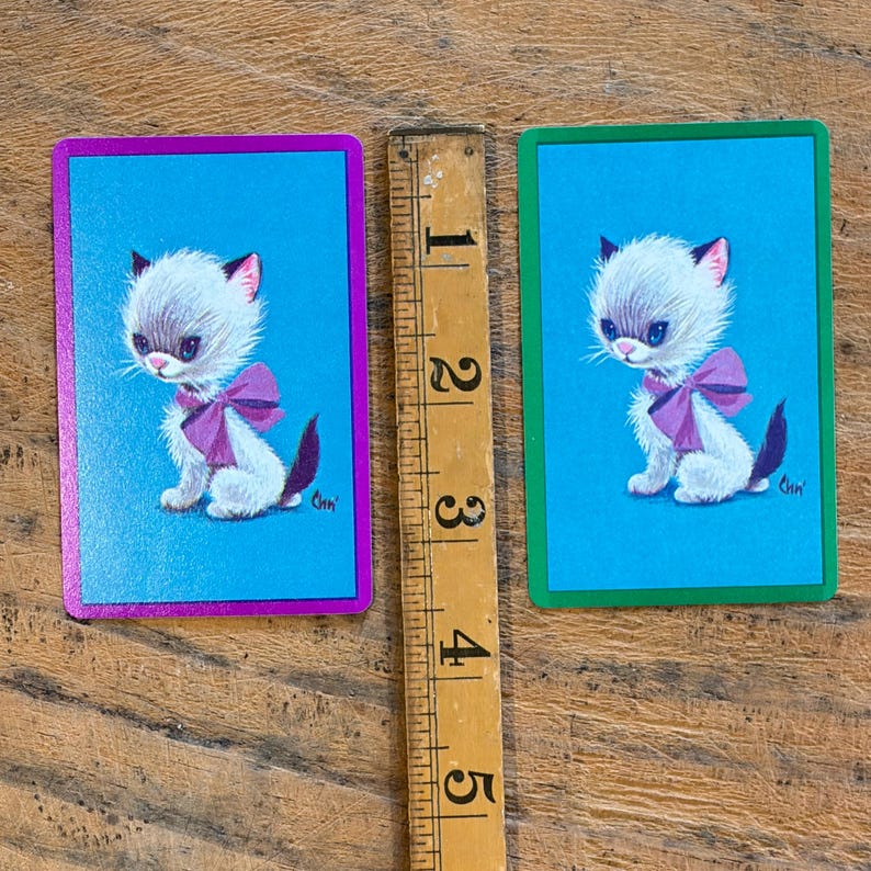 May include: Two rectangular playing cards featuring a white kitten with a purple bow on a bright blue background. One card has a purple border and the other a green border. A wooden ruler is placed between them, showing a height of 3.5 inches.