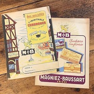 May include: Two vintage advertisements for a chocolate and confectionery shop called "Magnier-Baussart" in Amiens, France. The top ad features a cartoon illustration of a building with a cat and the text "L'Inimitable Caranouga Le Seul... Le Vrai... Tom-Cat Goffées assortis MOB". The bottom ad features a red and white chocolate bar with the text "Chocolat Heralda Magnier-Baussart A Croquer" and a smaller chocolate bar with the text "Chocolat Fourie Magnier-Baussart Creme Framboise".