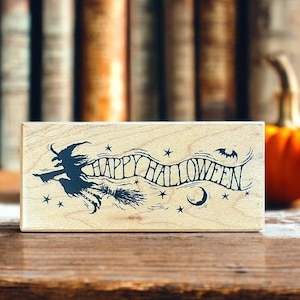 May include: A wooden stamp with the words "HAPPY HALLOWEEN" in a decorative font. The design includes a witch on a broom, stars, a crescent moon, and bats, all in black ink. The stamp is displayed on a wooden surface.