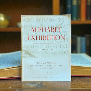 May include: A vintage book titled "Alphabet Exhibition" with red lettering on a cream-coloured cover. The book, published in London in 1953, features text about the alphabet throughout history. The book is displayed in front of a bookshelf.