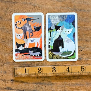 May include: Two rectangular cards featuring cat illustrations next to a wooden ruler showing 5 inches. The left card displays stacked cats in orange and grey tones. The right card shows two cats against a blue floral background with white borders.