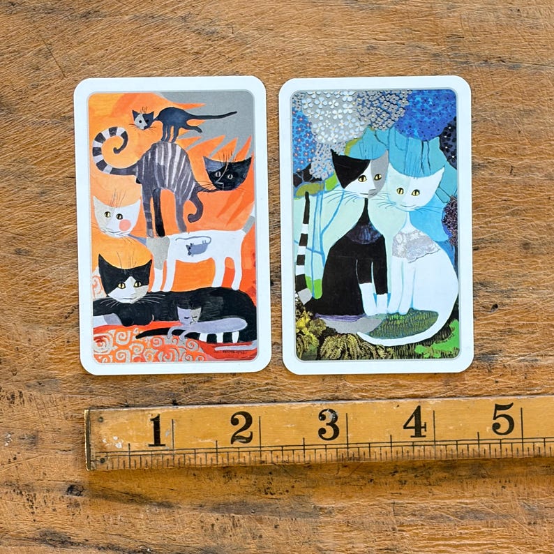 May include: Two rectangular cards featuring cat illustrations next to a wooden ruler showing 5 inches. The left card displays stacked cats in orange and grey tones. The right card shows two cats against a blue floral background with white borders.