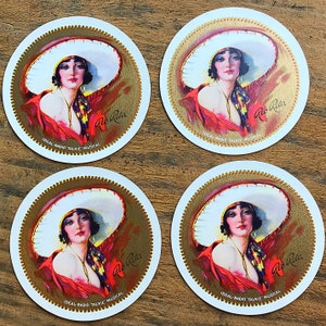 May include: Four round coasters featuring a vintage illustration of a woman in a large white hat and red shawl. The coasters have a gold border and the text "Rio Rita" and "Ideal-Radio Talkie Musical".