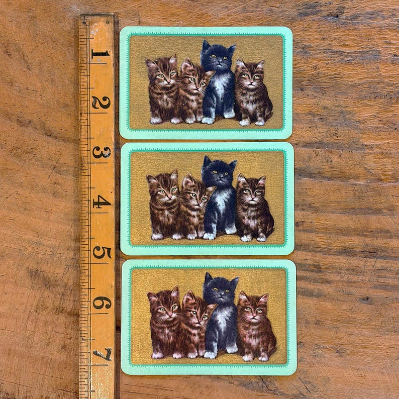 May include: Three vintage playing cards featuring illustrations of kittens. Each card has a gold background and a light green border. The kittens are depicted in various poses, with colors ranging from brown to black.