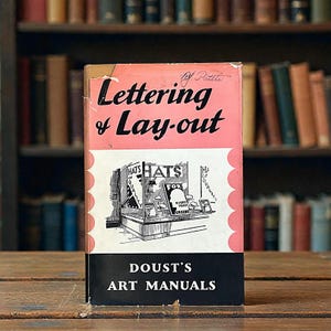 May include: A vintage book titled "Lettering & Lay-out" by Patite. The cover is pink and black with the title in large black letters. A black and white illustration of a hat shop is featured. The book is part of "Dousts Art Manuals" series.