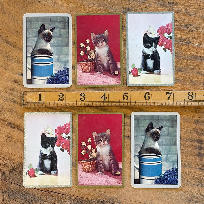 May include: Six vintage-style playing cards featuring cat illustrations. The cards showcase various cat breeds, including Siamese and tabby cats, in charming poses. Some cards include floral accents and a blue and white mug. The cards are arranged on a wooden surface.