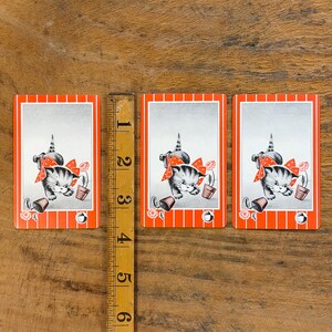 May include: Three vintage playing cards featuring a gray and white kitten wearing a red bow. The kitten is next to a flower pot and a rose. The cards have a red and white striped border and a silver background. A wooden ruler is next to the cards.