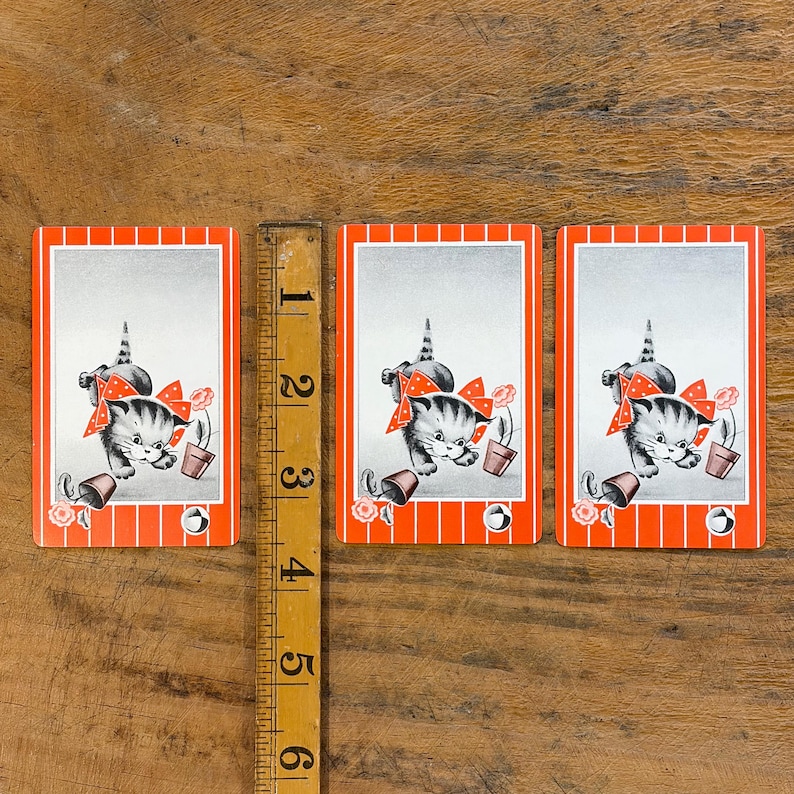 May include: Three vintage playing cards featuring a gray and white kitten wearing a red bow. The kitten is next to a flower pot and a rose. The cards have a red and white striped border and a silver background. A wooden ruler is next to the cards.