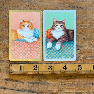May include: Two rectangular playing cards featuring illustrations of cats with yarn balls, positioned above a wooden ruler showing 5 inches. The left card has a ginger cat on a peach background, and the right card has a brown tabby cat on a mint green background.