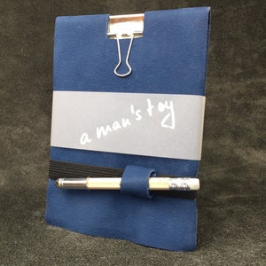 Pode incluir: Um caderno de couro azul com um clipe de metal prateado e uma banda elástica preta. O caderno tem uma etiqueta branca com o texto "a man's toy" escrito nela. Um lápis está preso ao caderno com a banda elástica.