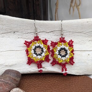 May include: A pair of beaded earrings with a starburst design. The earrings feature red, yellow, white, and black beads. The earrings are suspended from silver-colored fish hook ear wires. The earrings are displayed on a piece of driftwood.