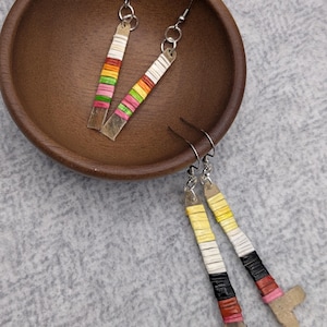 May include: Two pairs of dangle earrings featuring long, thin, flat rectangular components wrapped in colorful thread. One pair displays bands of white, orange, yellow, green, and pink. The other pair features bands of yellow, white, black, and red.