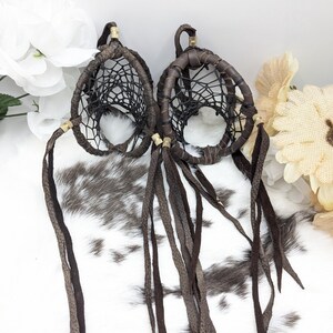 May include: Two brown leather dream catchers with intricate woven webbing. Each dream catcher has long, dangling leather strips and gold-colored accents. The dream catchers are displayed against a white and brown spotted background, with white and beige flowers.