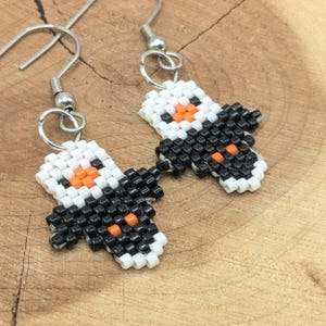 May include: A pair of dangle earrings featuring a beaded penguin design. Each earring is crafted from small white, black, and orange seed beads arranged to form a penguin shape, suspended from silver-toned metal fishhook ear wires.