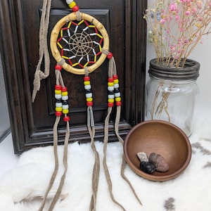 May include: A handcrafted dreamcatcher with a circular wooden frame, featuring a web design in black, red, and white. The dreamcatcher has beaded accents in red, yellow, black, and white, and long leather strands. The background includes a wooden frame, a wooden bowl, and a jar with flowers.