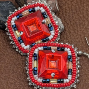 May include: A pair of square-shaped earrings with a central red, faceted gem. The earrings are framed with red, white, blue, and gold beads, and have silver-colored ear wires. The design is geometric and eye-catching.