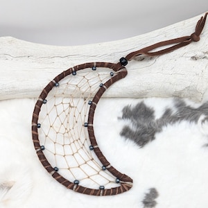 May include: Crescent moon dreamcatcher with a brown suede-wrapped frame and a woven web. The dreamcatcher is adorned with small, dark blue beads and a brown leather hanging cord. The background features a white fur-like texture and a piece of driftwood.