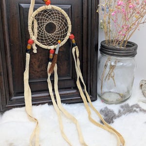 May include: A cream-colored dreamcatcher with a woven web design and dangling leather strips. The dreamcatcher is adorned with red, black, and brown beads. A small, decorative feather is attached to the center. The dreamcatcher is a decorative item.