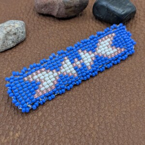 May include: A beaded barrette with a blue background and white and pink star-like designs. The barrette is rectangular and made of small, colorful beads. The barrette is on a brown surface with three small stones.