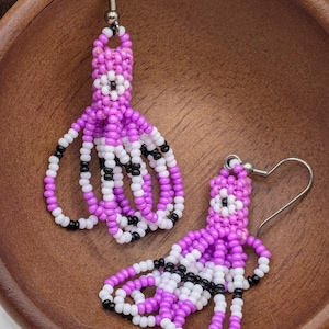 May include: Handmade beaded earrings in a unique octopus design. The earrings feature a combination of purple, white, and black beads. The octopus's head is a solid purple, with white and black beads forming the eyes. The tentacles are made of alternating white and black beads.