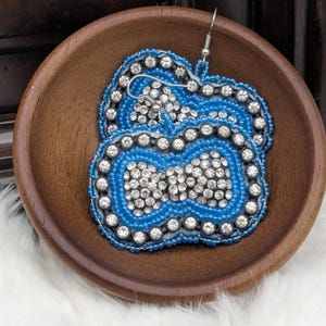 May include: A pair of blue beaded earrings with a central design of sparkling rhinestones. The earrings are set in a wooden bowl on a white, fluffy surface. The earrings have a silver hook.