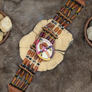 May include: Handmade beaded choker necklace with a peace sign pendant. The necklace features brown wooden beads, black beads, and leather accents. The pendant is round with a colorful peace sign design.
