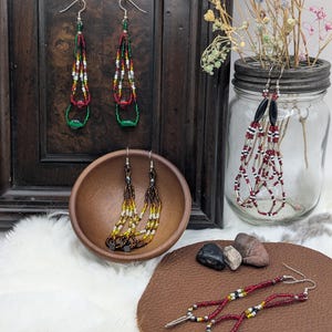 May include: Various beaded earrings in different styles. Some are displayed on a wooden frame, others in a wooden bowl, and some on a leather surface. The earrings feature a variety of colors, including red, green, yellow, and black, with silver-tone hooks.