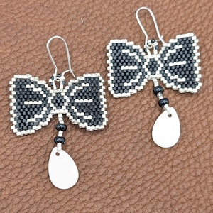 May include: A pair of handmade bow-tie earrings. The earrings feature a black and white beaded design, with a silver-colored metal bow shape. Each earring has a small cluster of black beads and a white teardrop-shaped charm. The earrings hang from silver-colored hooks.