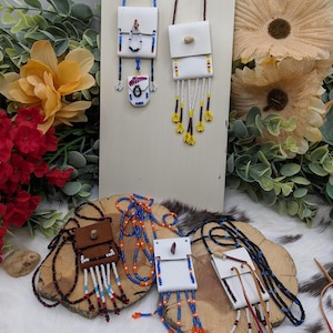 May include: Handmade leather pouch necklaces with beaded straps. The pouches are white, brown, and off-white, with beaded fringe and accents in red, blue, orange, and yellow. The necklaces are displayed on wooden slices and a white background.