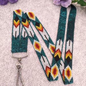 May include: A beaded lanyard featuring a dark teal background with a repeating pattern of white, yellow, orange, and black feather-like shapes. The strap is finished with a silver-toned metal swivel hook clasp at the bottom for attaching items.