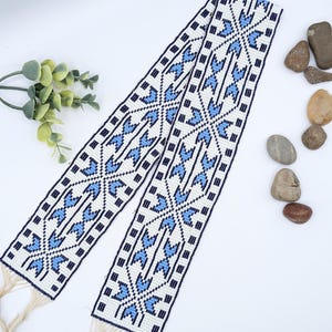 May include: A beaded belt with a white background, featuring a geometric pattern in blue and black. The belt has a fringe at the ends. The design includes blue arrow-like shapes and black squares, creating a symmetrical pattern.