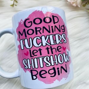 May include: White ceramic mug with a pink watercolour-style background. The mug features the text "Good Morning Fuckers Let the Shitshow Begin" in black, with pink hearts. The mug has a white handle.