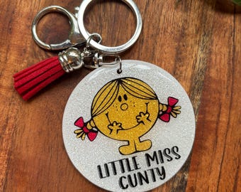 Puede incluir: Un llavero de purpurina blanca con un personaje de dibujos animados amarillo con cabello rojo y un lazo rojo. El texto "Little Miss Cunty" está impreso debajo del personaje.