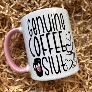 May include: White ceramic coffee mug with a pink handle. The mug features the words "Genuine COFFEE Slut" in black script, along with coffee-themed illustrations and hearts. The mug is set against a background of brown paper.