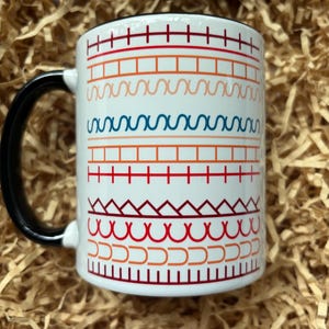 May include: White ceramic mug with a black handle and rim. The mug features a repeating pattern of colourful geometric shapes in red, orange, and blue. The mug is sitting on a bed of brown packing material.