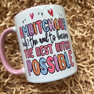 May include: White ceramic mug with a pink handle and rim. The mug features the text "AMBITCHOUS (adj.) the need to become THE BEST BITCH POSSIBLE" in colourful bubble letters. Decorative hearts and stars are also present.