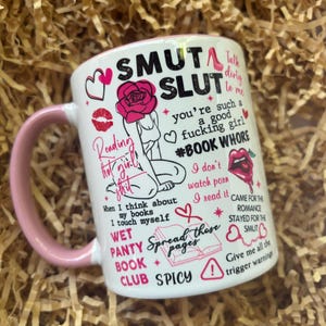 May include: White ceramic mug with a pink handle. The mug features a pink and black design with the text "SMUT SLUT" and other phrases. The mug is sitting on a bed of brown crinkled paper.