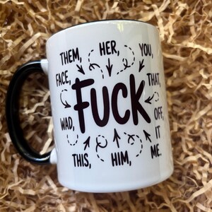 May include: White ceramic mug with a black handle and rim. The mug features the word "FUCK" in large black letters, surrounded by other words and phrases like "THEM", "HER", "YOU", "THIS", and "HIM".