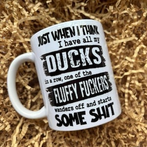 May include: White ceramic mug with a black humorous quote: "JUST WHEN I THINK I have all my DUCKS in a row, one of the FLUFFY FUCKERS wanders off and starts SOME SHIT." The mug has a curved handle and is set against a brown background.