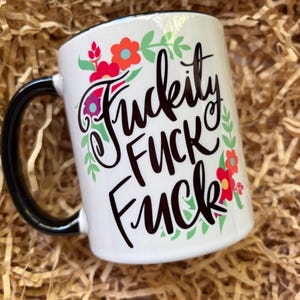 May include: White ceramic mug with a black handle and rim. The mug features the words "Fuckity Fuck Fuck" in black script, surrounded by floral and leaf designs in red, orange, and green.
