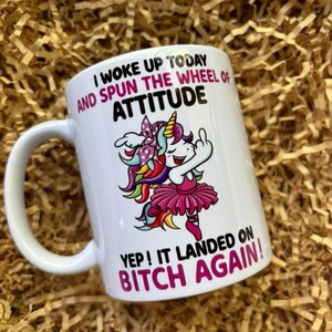 May include: White ceramic mug featuring a unicorn graphic with a rainbow mane and pink tutu. The mug has the text "I WOKE UP TODAY AND SPUN THE WHEEL OF ATTITUDE. YEP! IT LANDED ON BITCH AGAIN!"