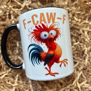 May include: White ceramic mug with a black handle and rim. The mug features a cartoon rooster illustration with orange text that reads "F-CAW-F". The rooster is red, orange, and blue with wide eyes.